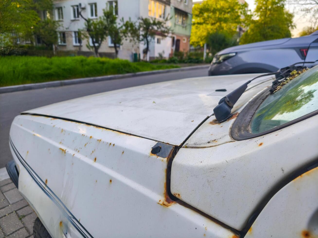 A white car with a rusted hood and a dirty windshield. The car is parked on a street next to a building photo