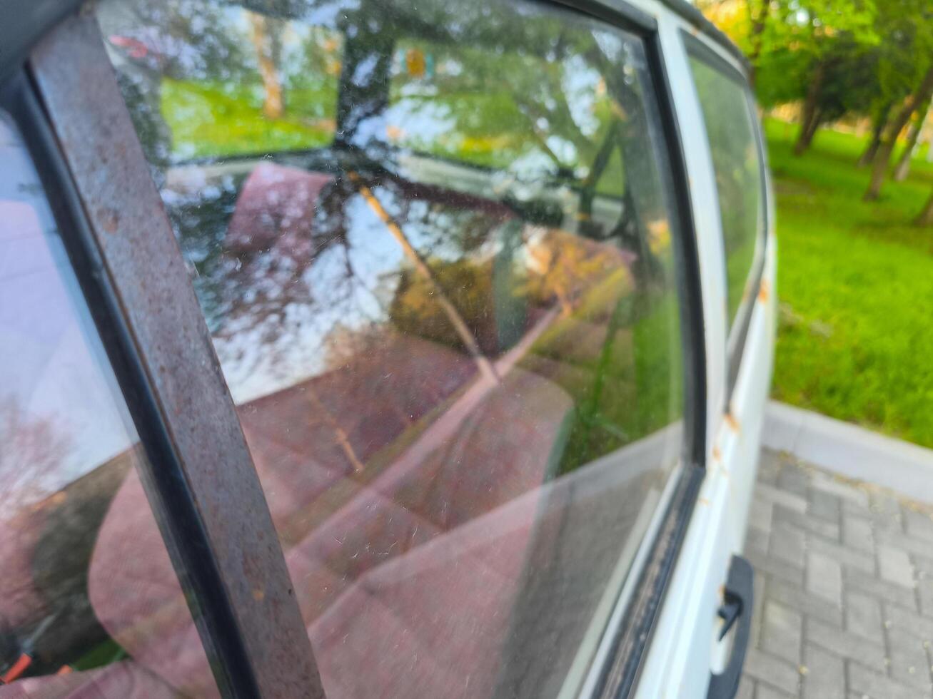 A car window is dirty and has a reflection of trees in it. The car is parked in a grassy area photo