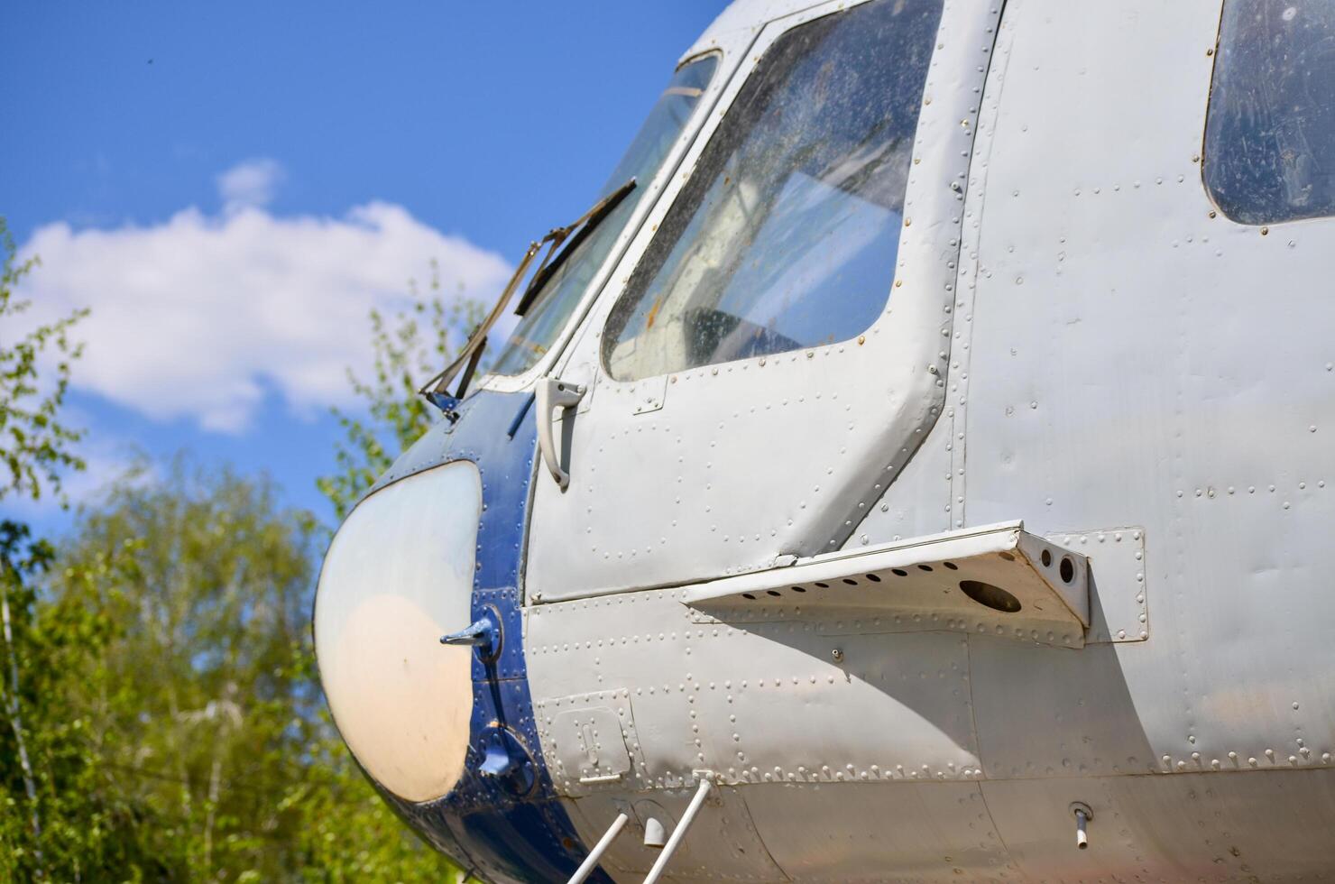 A small plane with a blue and white paint job sits on the ground. The plane is parked in a wooded area, with trees surrounding it. The sky is clear and blue, and the sun is shining brightly photo