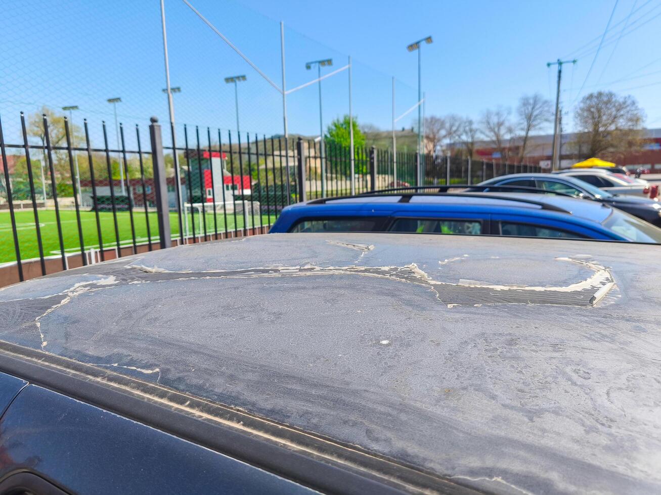 A car has a large mark on the hood, and the hood is covered in dirt. The car is parked in a lot with other cars, and there is a fence in the background. The scene gives off a sense of neglect photo