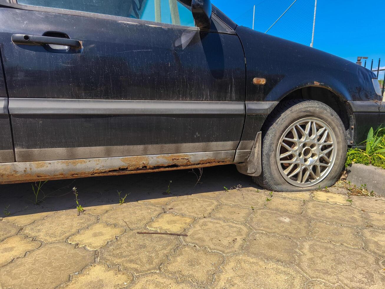 A dirty car with a tire that is flat. The car is parked on a brick road. The car is old and has a lot of dirt on it photo