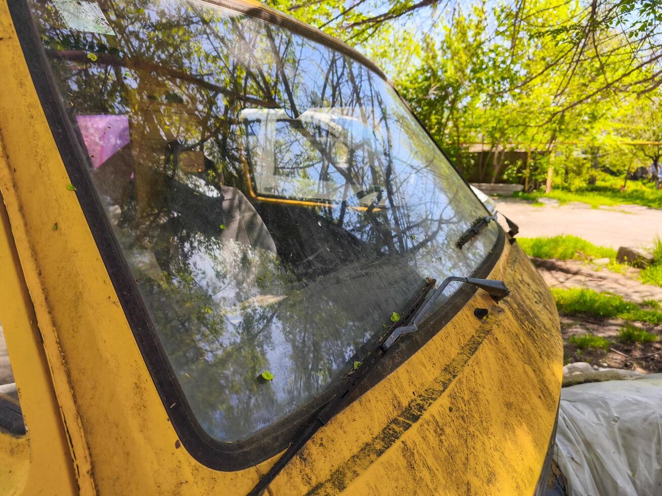 A yellow car with a dirty windshield and wipers. The windshield is covered in dirt and the wipers are covered in mud. The car is parked in a grassy area photo