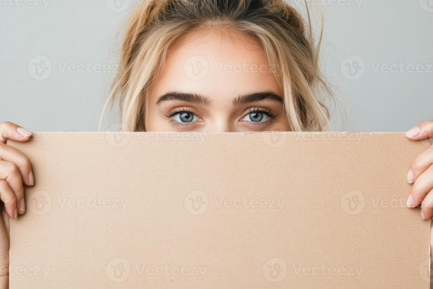 A young woman with blue eyes holds a blank cardboard sign in front of her face against a light background photo