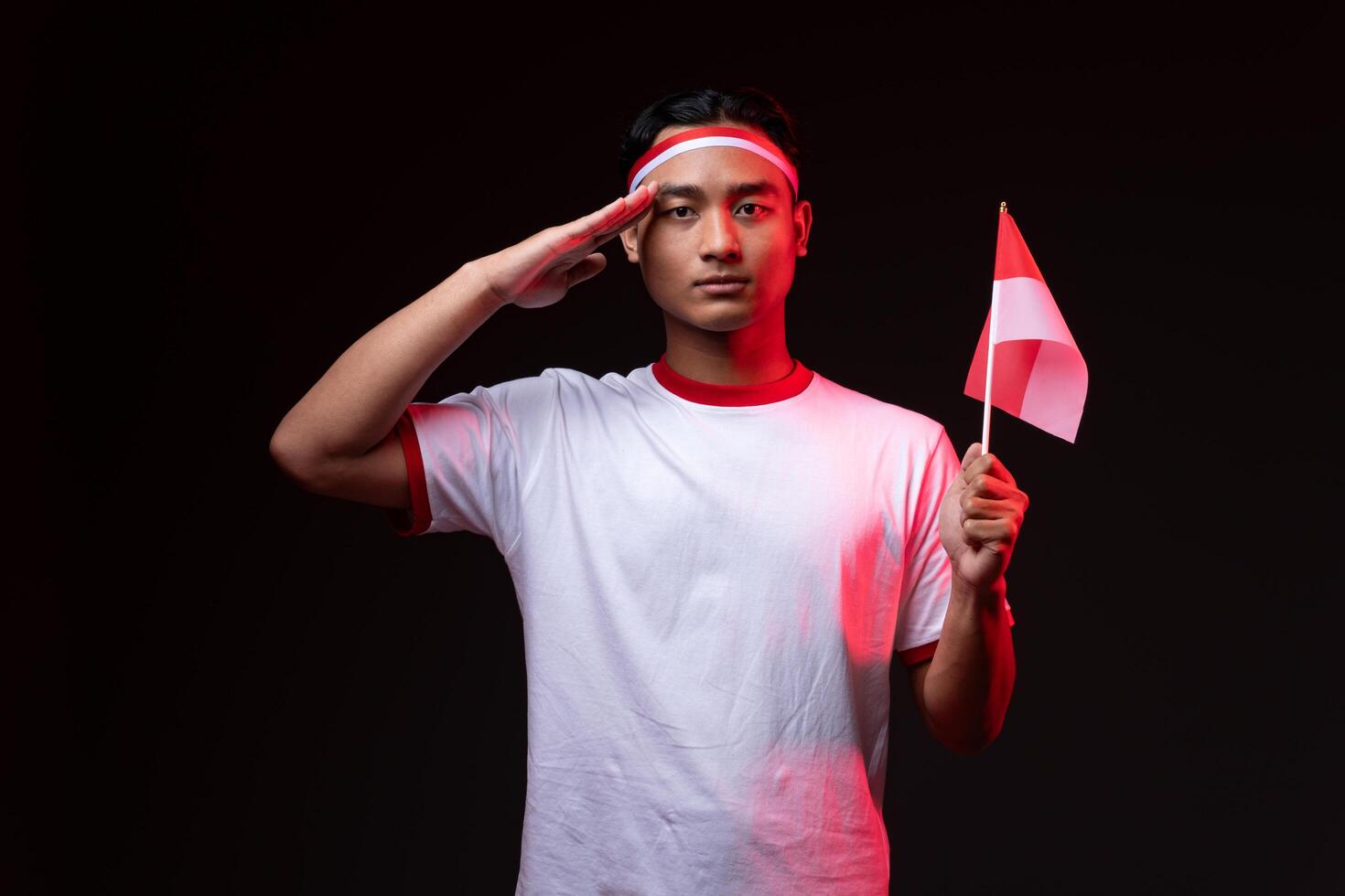 Portrait of Young Asian Man with Salute Gesture Wearing Red White Outfit, Indonesian Independence Day Concept Serious Isolated Flag photo