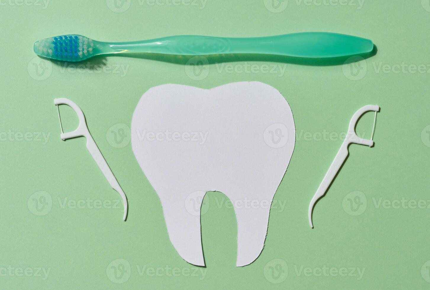 A green toothbrush, a white paper cutout of a tooth, and two dental floss picks arranged on a light green background photo