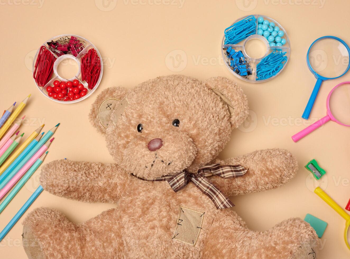 brown teddy bear is surrounded by various colorful school supplies on a neutral background photo
