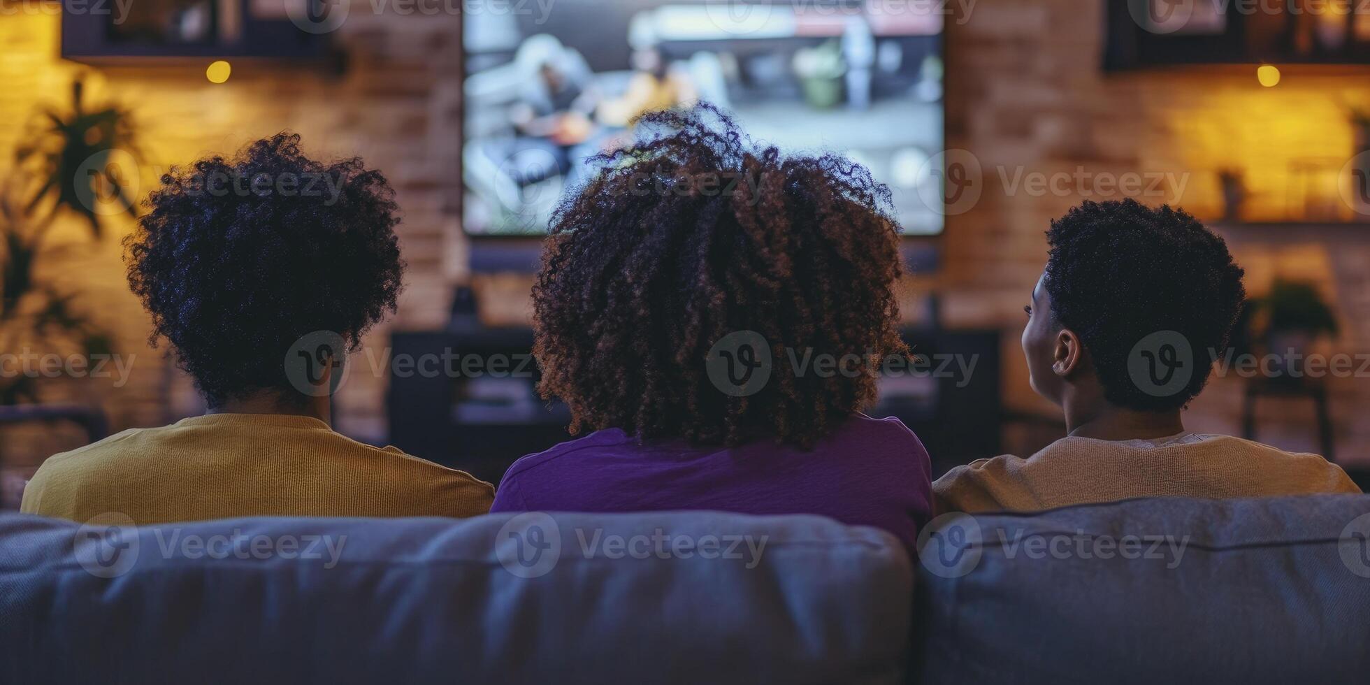 People are sitting on the couch watching TV. One of them is Black with curly hair and wearing a purple shirt. They look happy. photo