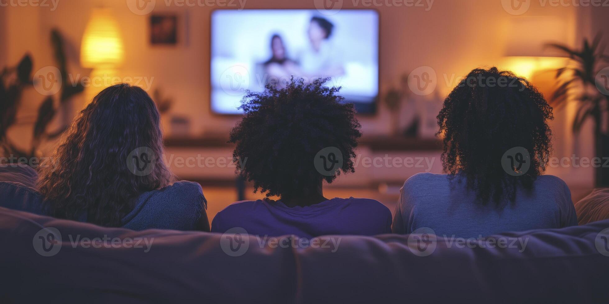 People are sitting on the couch watching TV. One of them is Black with curly hair and wearing a purple shirt. They look happy. photo