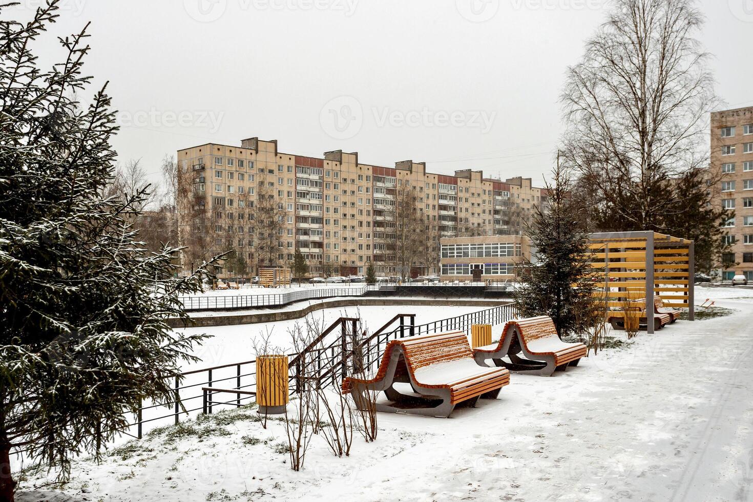 A snow-covered city park with benches and a fir tree against the backdrop of a typical Soviet residential building on a cloudy winter day. photo
