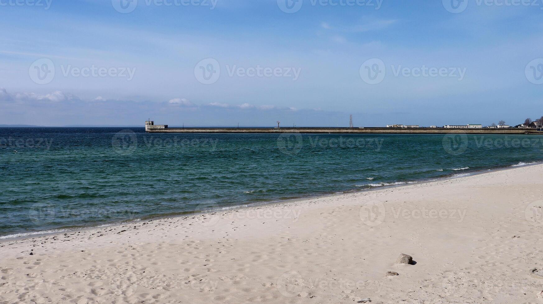Scenic view of a quiet sandy beach and calm sea waves under a clear blue sky. A breakwater structure with buildings and a watchtower stretches along the horizon in the distance. photo