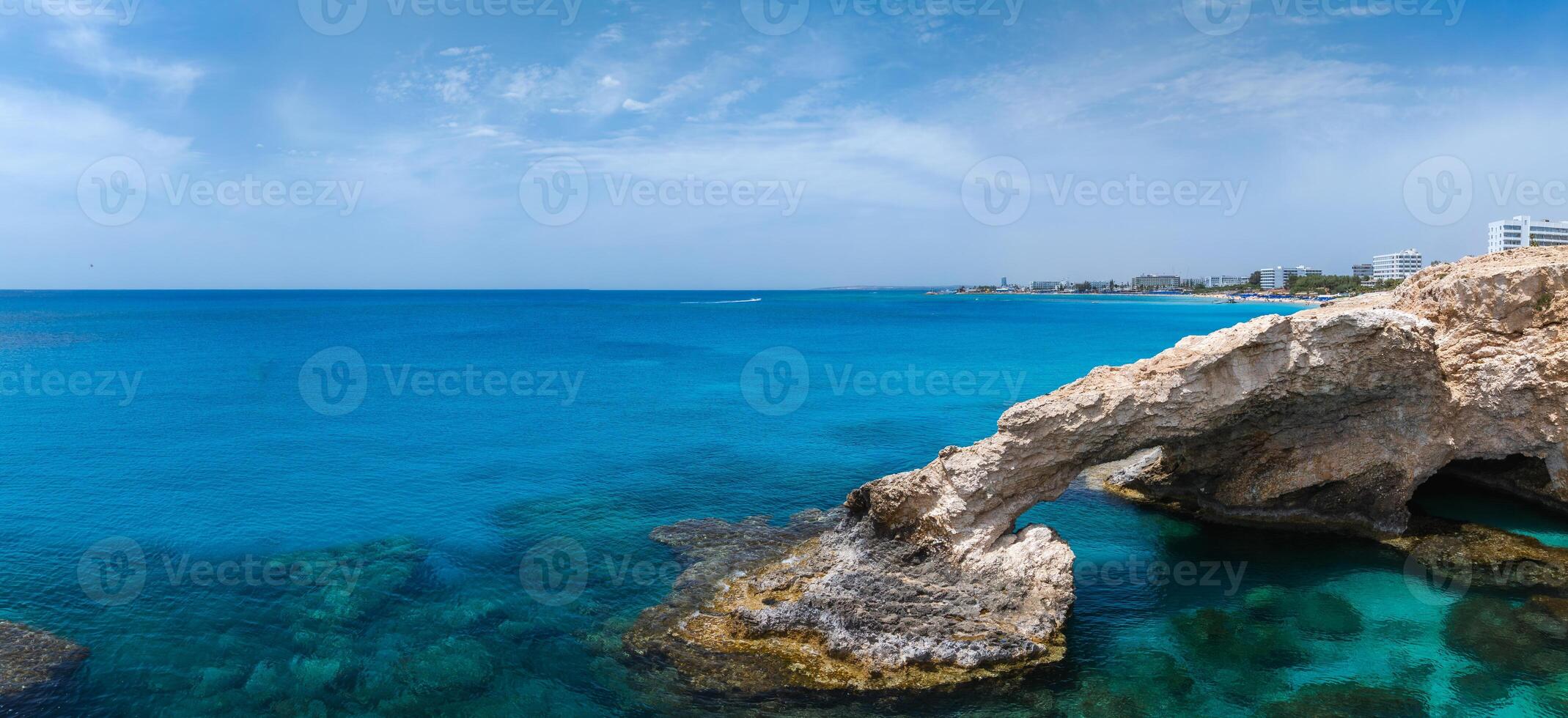 A natural Love arch spans over clear turquoise waters in Ayia Napa, Cyprus, with luxury hotels and resorts visible along the Mediterranean coastline. photo