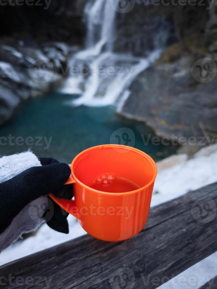 Gloved hand holding steaming orange mug near cascading waterfall, misty november landscape in aosta valley, italy Cascate di Lillaz photo