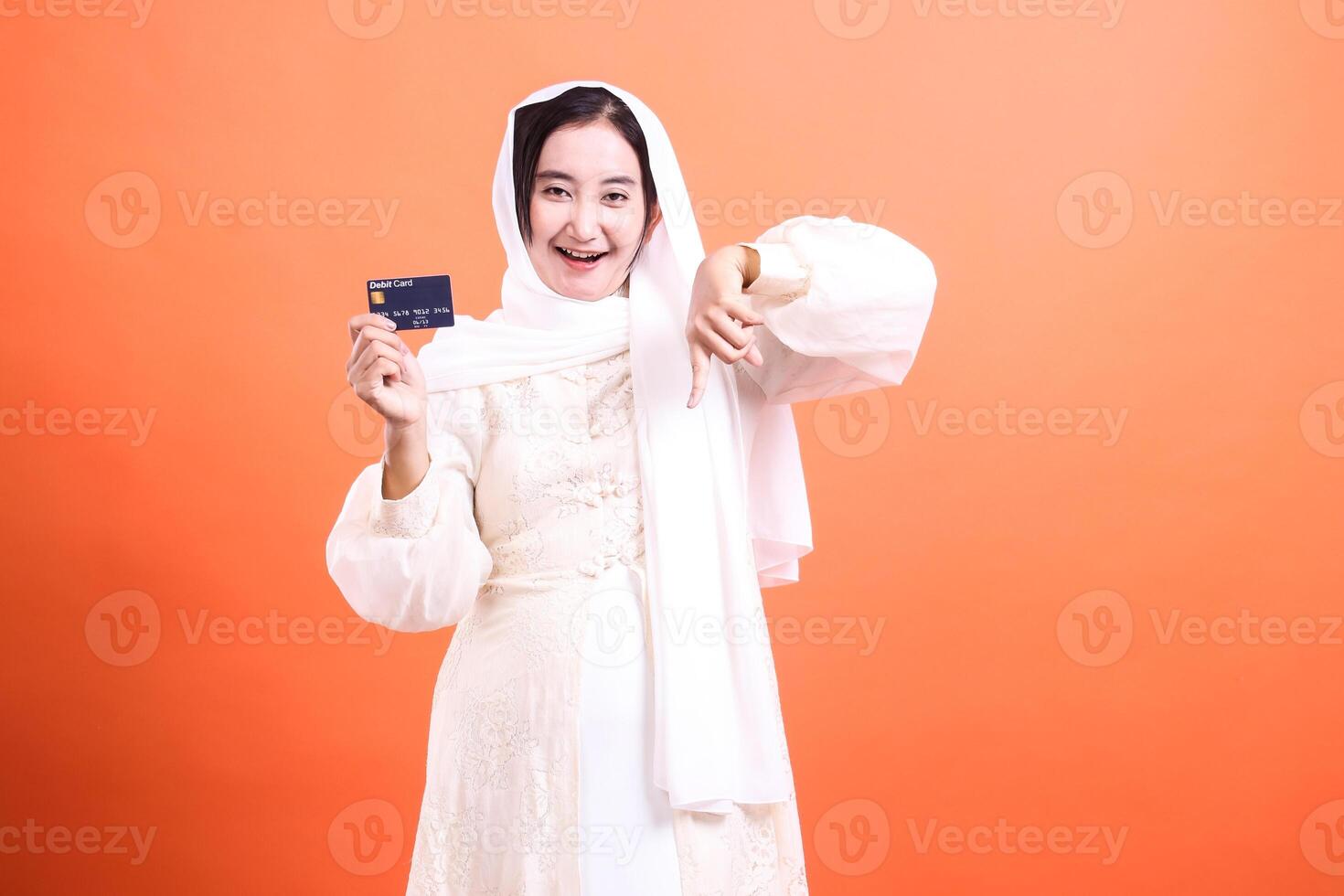Delighted Muslim Woman Presenting Credit Card with Enthusiasm in Front of Orange Background photo