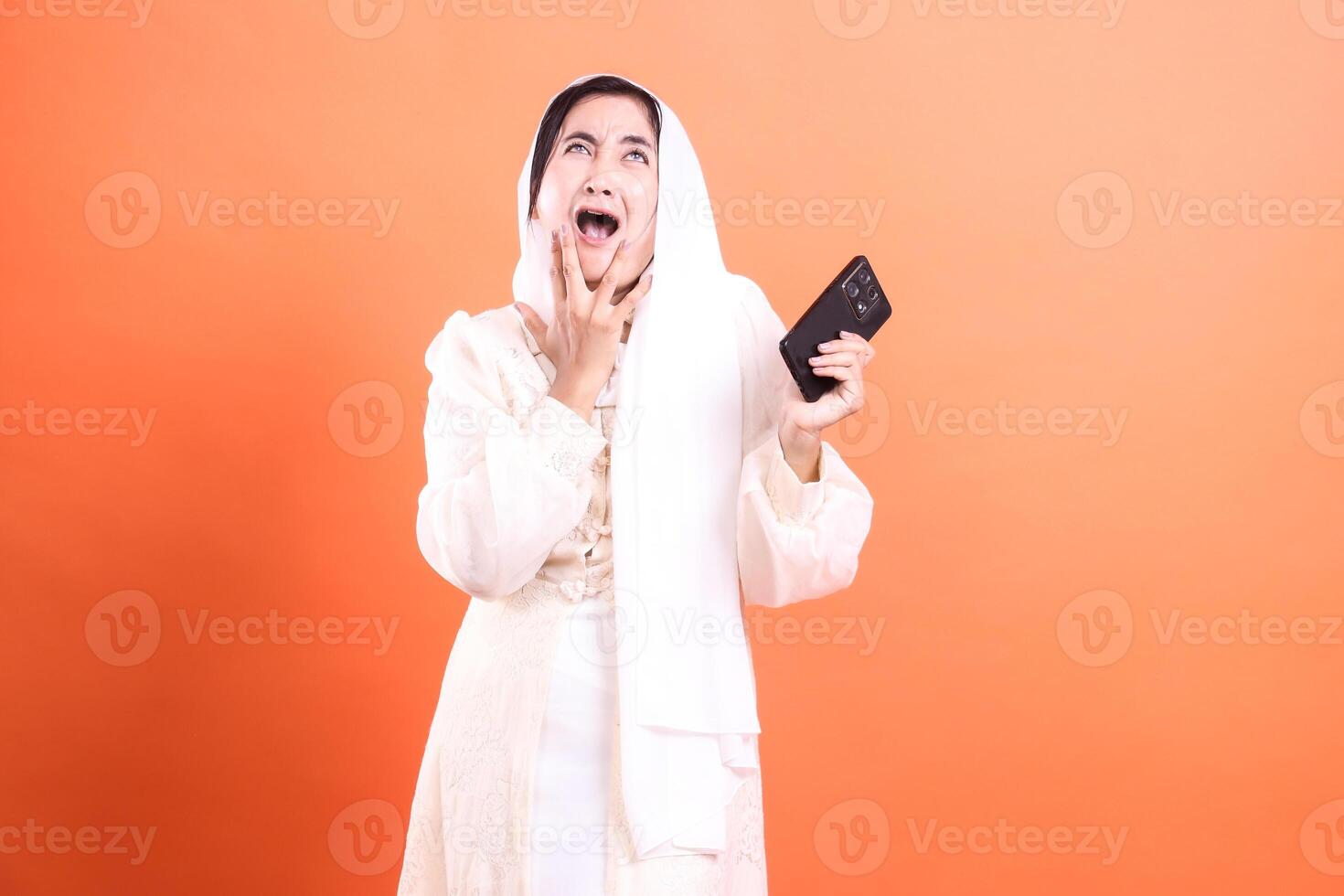 Shocked Muslim Woman with Smartphone A Moment of Disbelief and Surprise, isolated on a orange background, expressing intense emotion and disbelief. photo