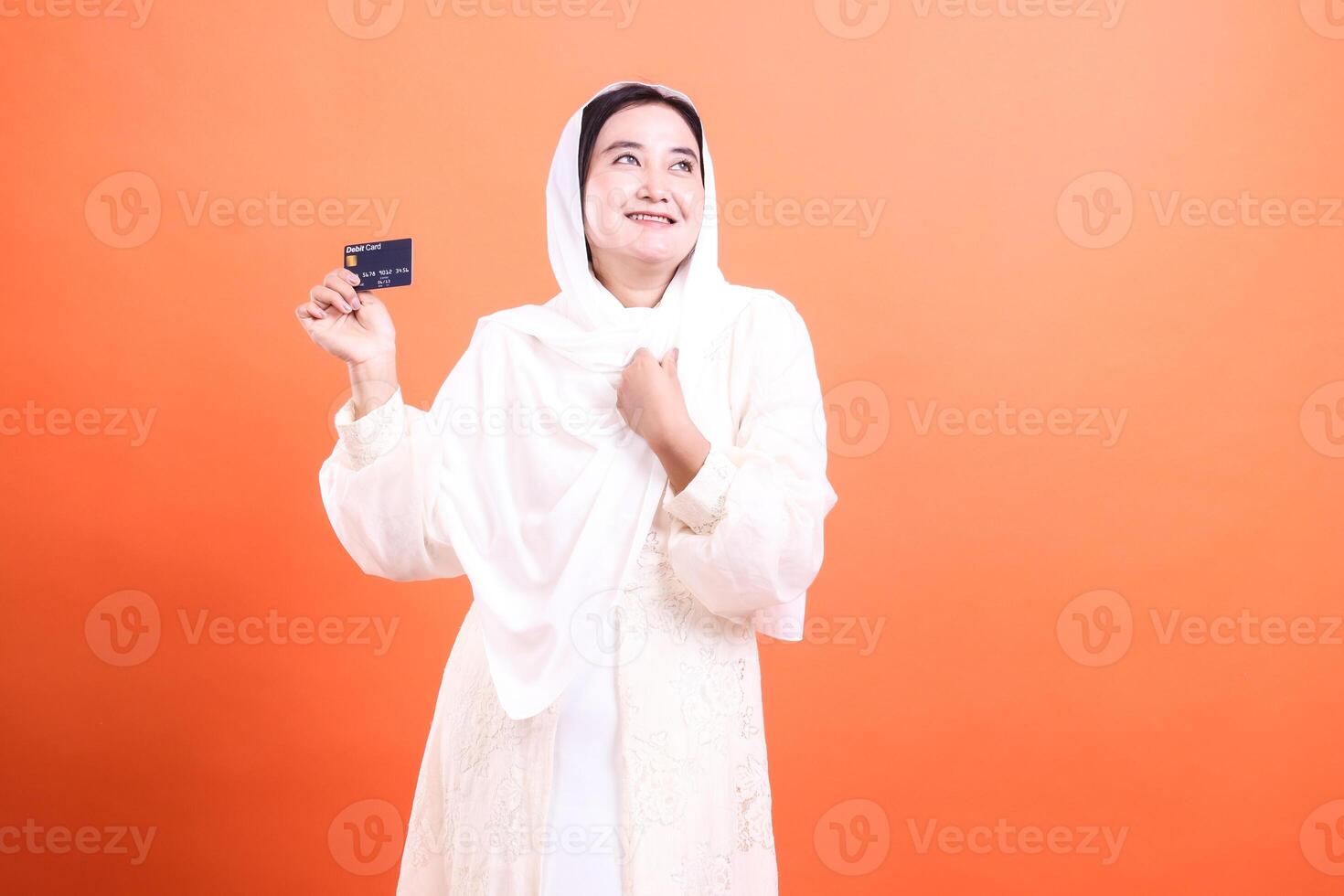 Happy Muslim Woman Holding Credit Card with a Smile, isolated on a orange background, symbolizing financial freedom and modern Islamic lifestyle. photo