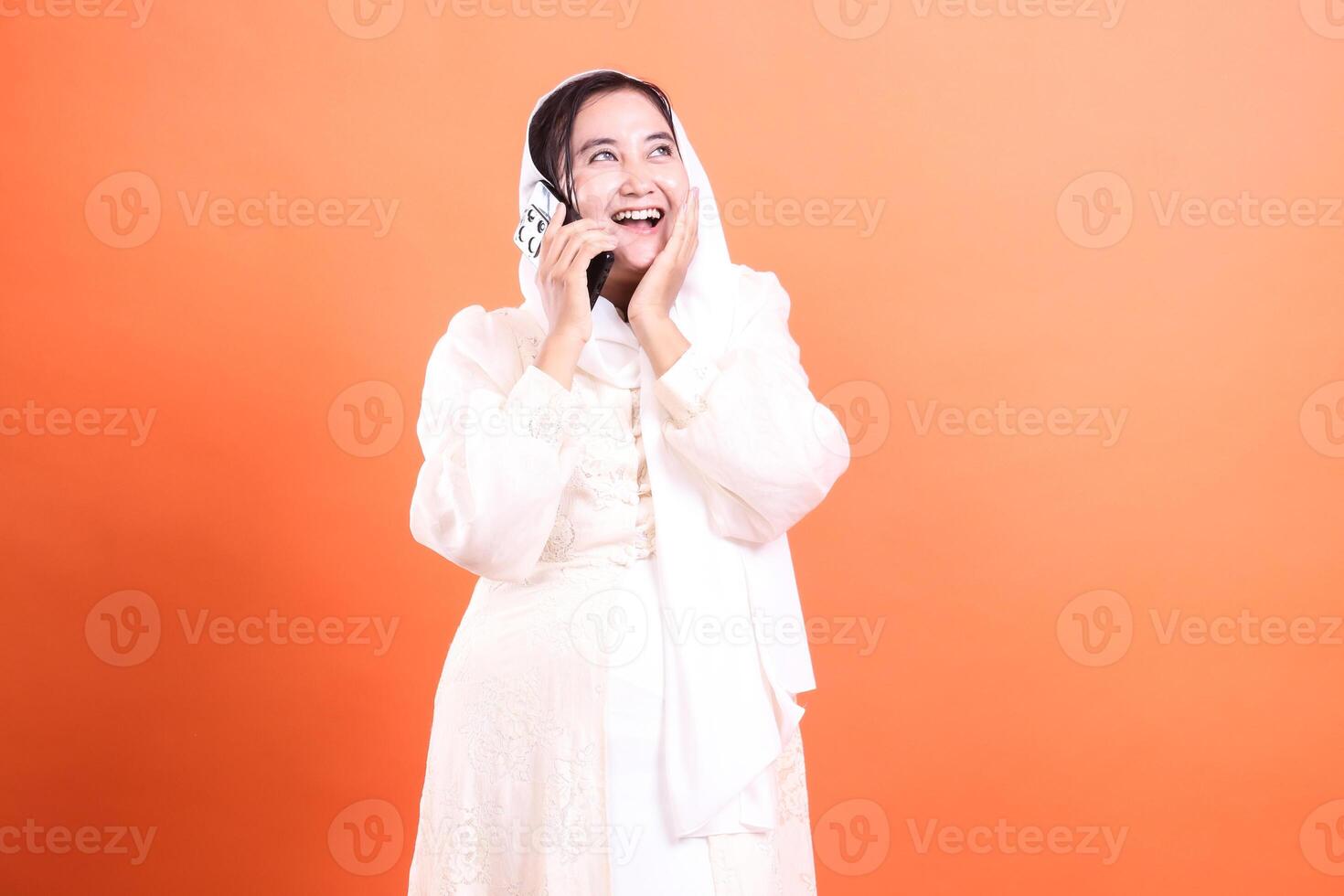 Joyful Muslim Woman Communicating A vibrant image capturing the modern Muslim woman enjoying a phone call with a smile, isolated on a orange background. photo