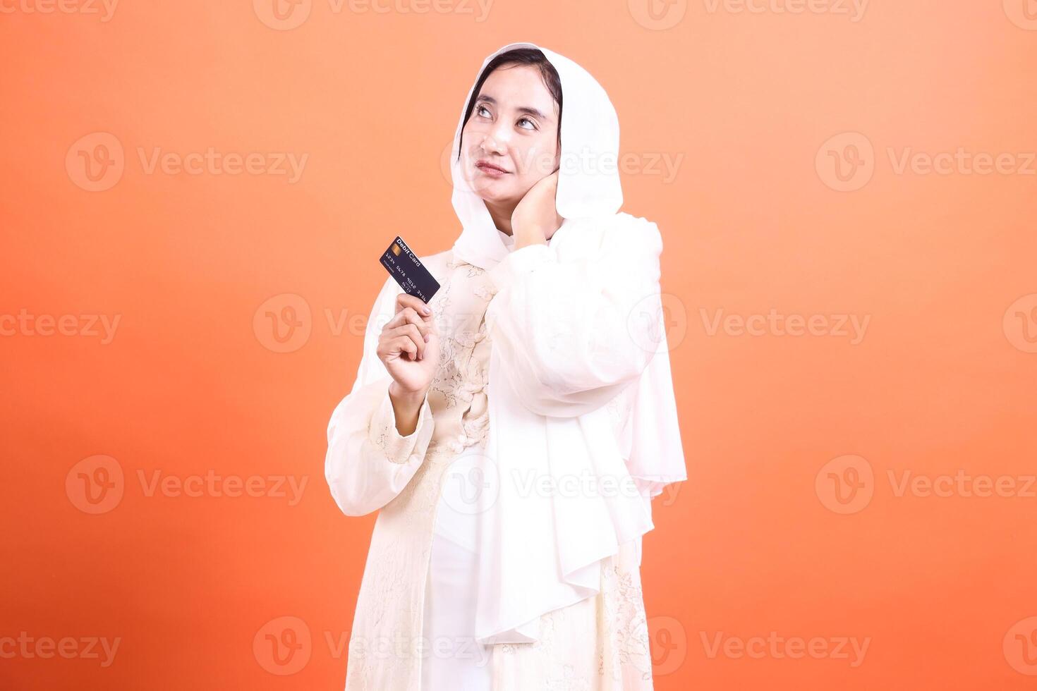 Young muslim woman thoughtfully holding debit card, looking up, contemplating online shopping possibilities, isolated on a orange background. photo