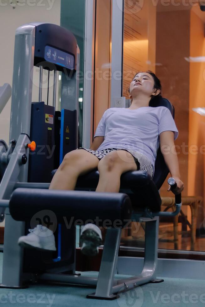 woman using leg extension machine at gym. sitting with legs positioned under padded machine bar wearing white shirt, short skirt, and white shoes photo