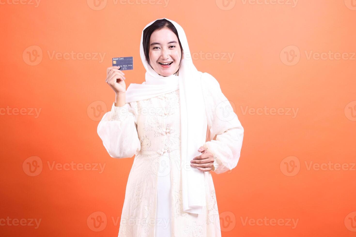 Happy Muslim Woman Showing Credit Card for Online Shopping, showcasing modern finance with a smile, isolated on a orange background. photo