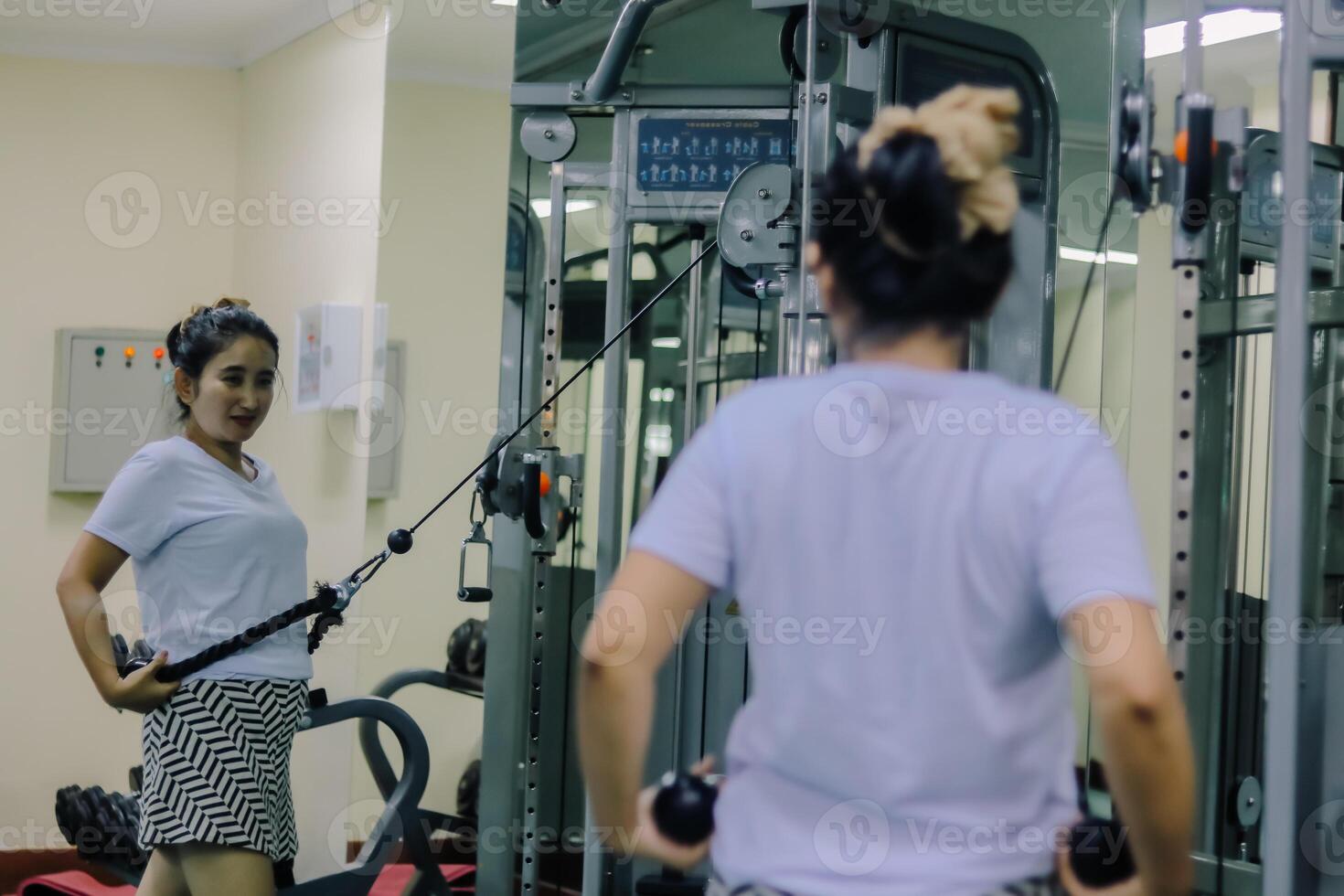A woman uses a cable exercise machine at a gym. She is facing the machine, but there is also a mirror reflection showing her side profile. photo