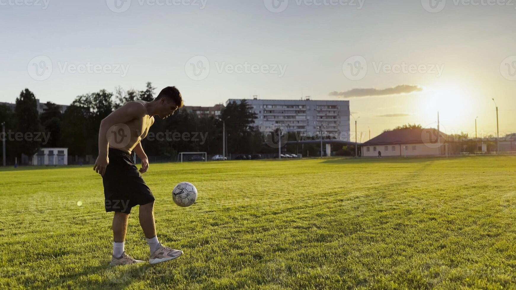 Young man juggling soccer ball on stadium at sunset. Professional footballer kicking ball at green field. Sportsman practicing tricks at meadow with sunlight at backgroud. Freestyle football photo