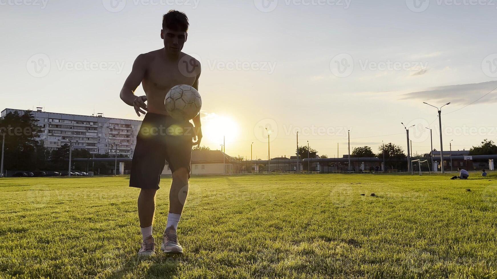 Young man juggling soccer ball on stadium at sunset. Professional footballer kicking ball at green field. Sportsman practicing tricks at meadow with sunlight at backgroun. Freestyle football photo