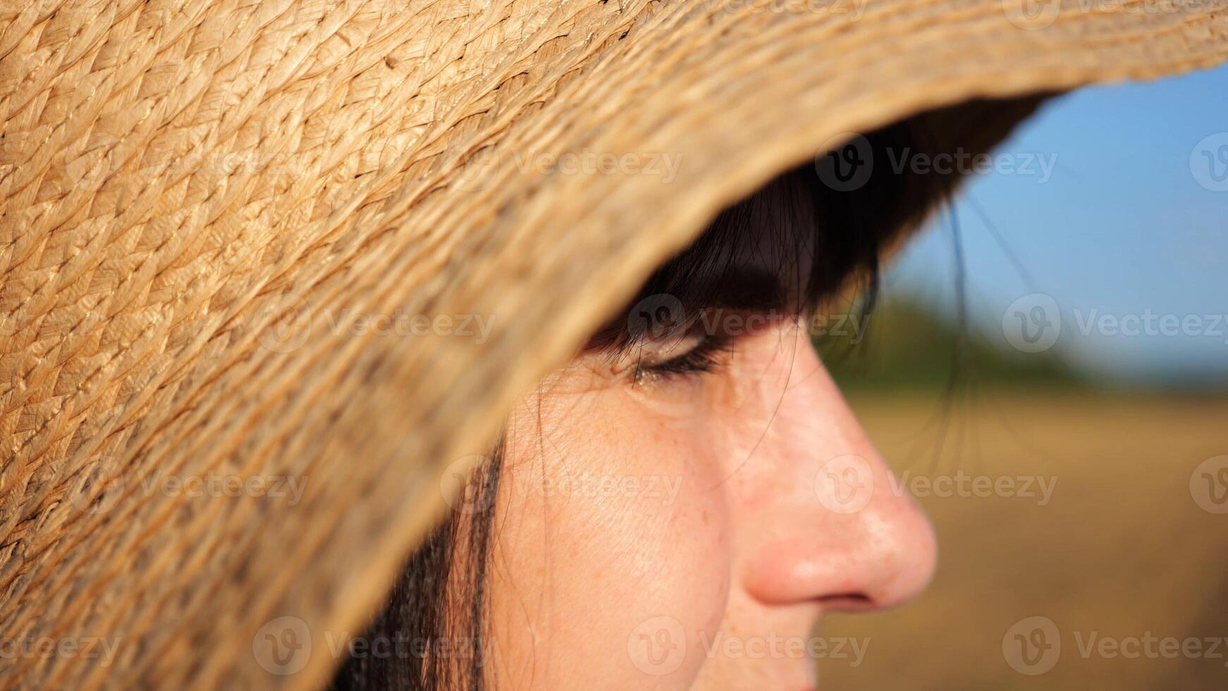 Female farmer in straw hat looking into wheat field and examining plantation at sunset. Close up face of adult beautiful woman illuminating by bright rays of sun. Concept of agriculture business. photo