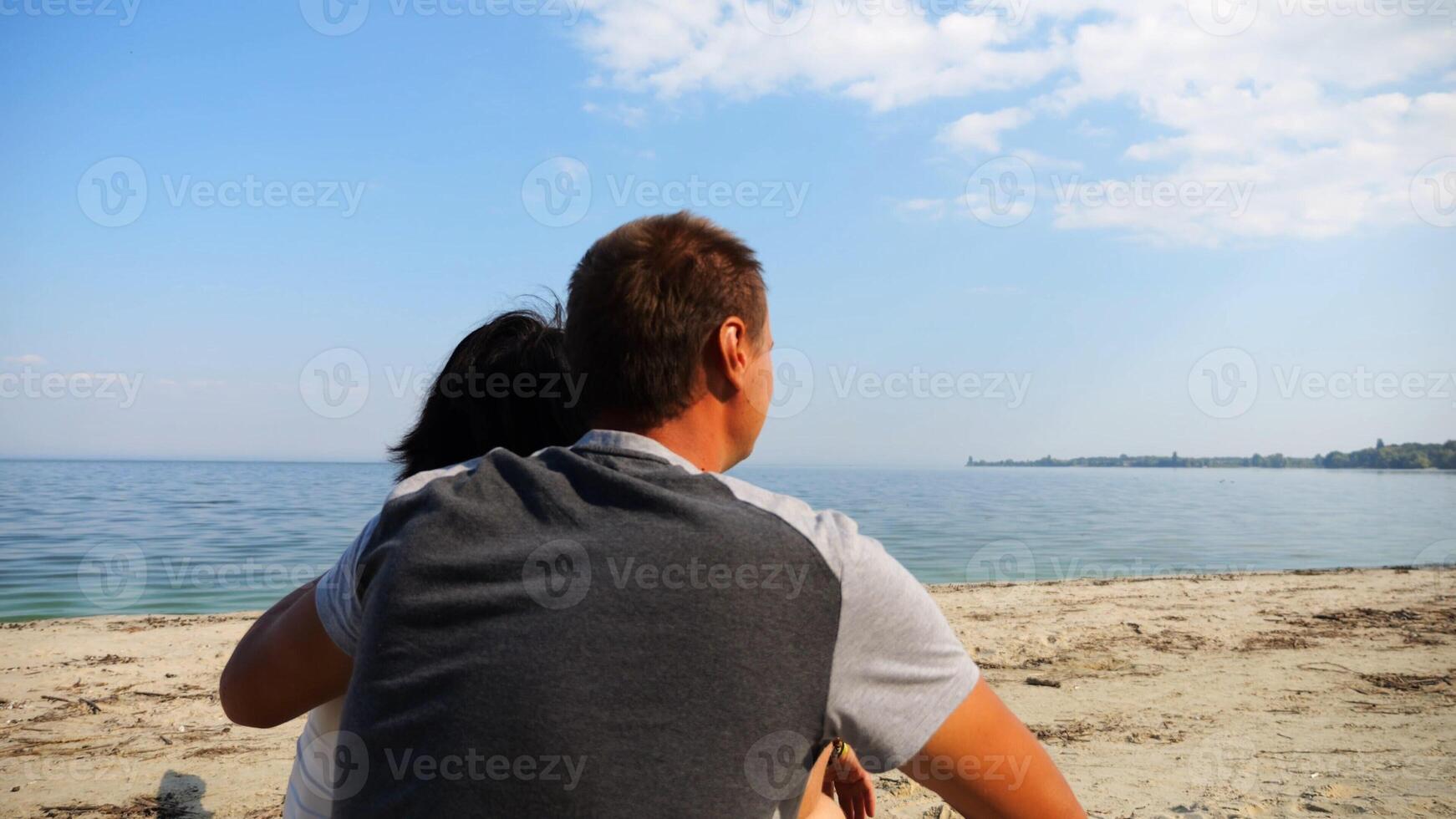 Happy pair hugs during sitting on wooden log on beach.Romantic couple admiring sea landscape at sunny day. Lovers spending time together at resort. Concept of summer vacation or holiday. Slow mo photo