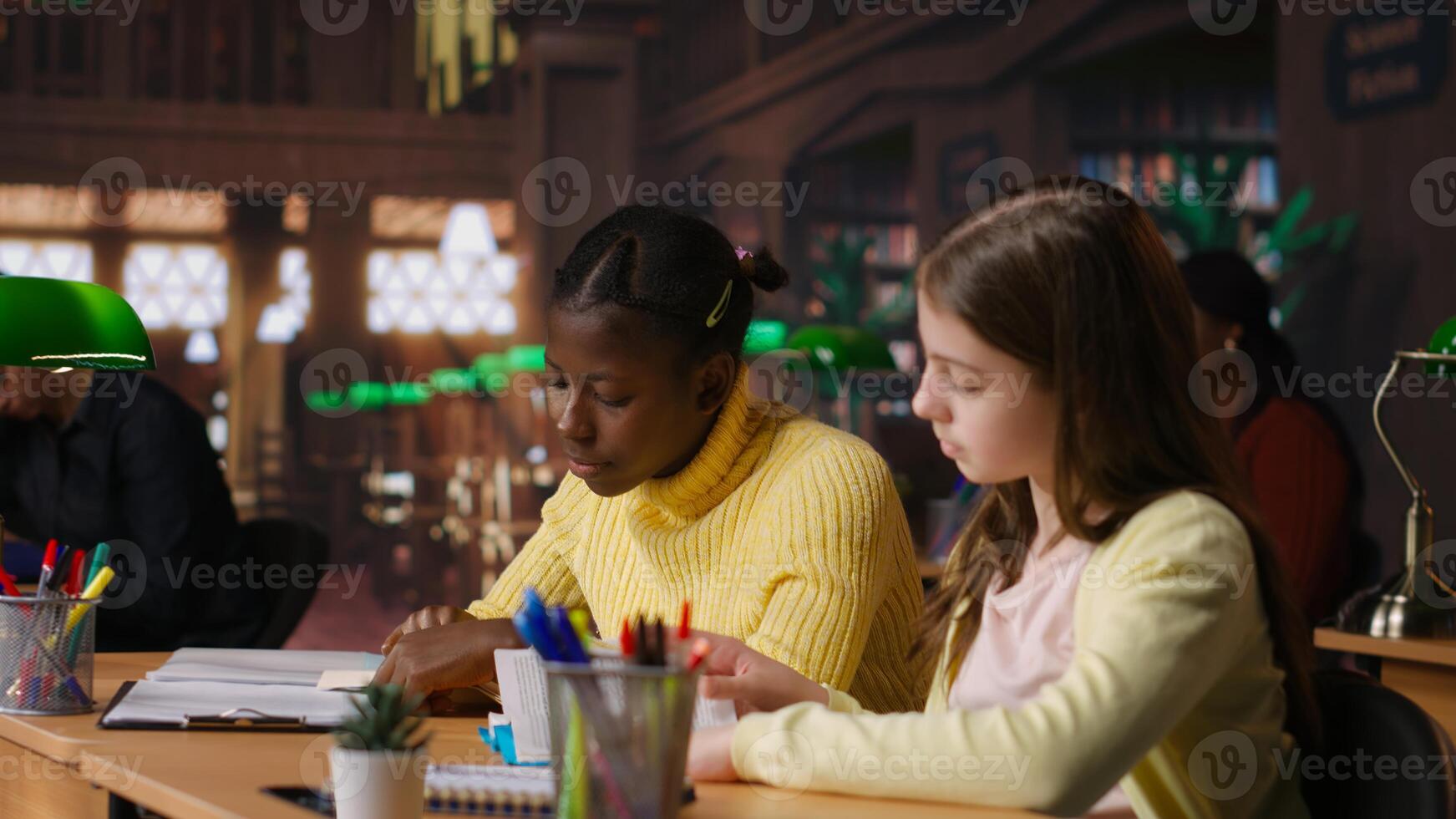 Preteen girls focus on reading required books at study desk, developing academic skills and literary language at library. Scholars enjoy lecture for school curriculum. Camera B. photo