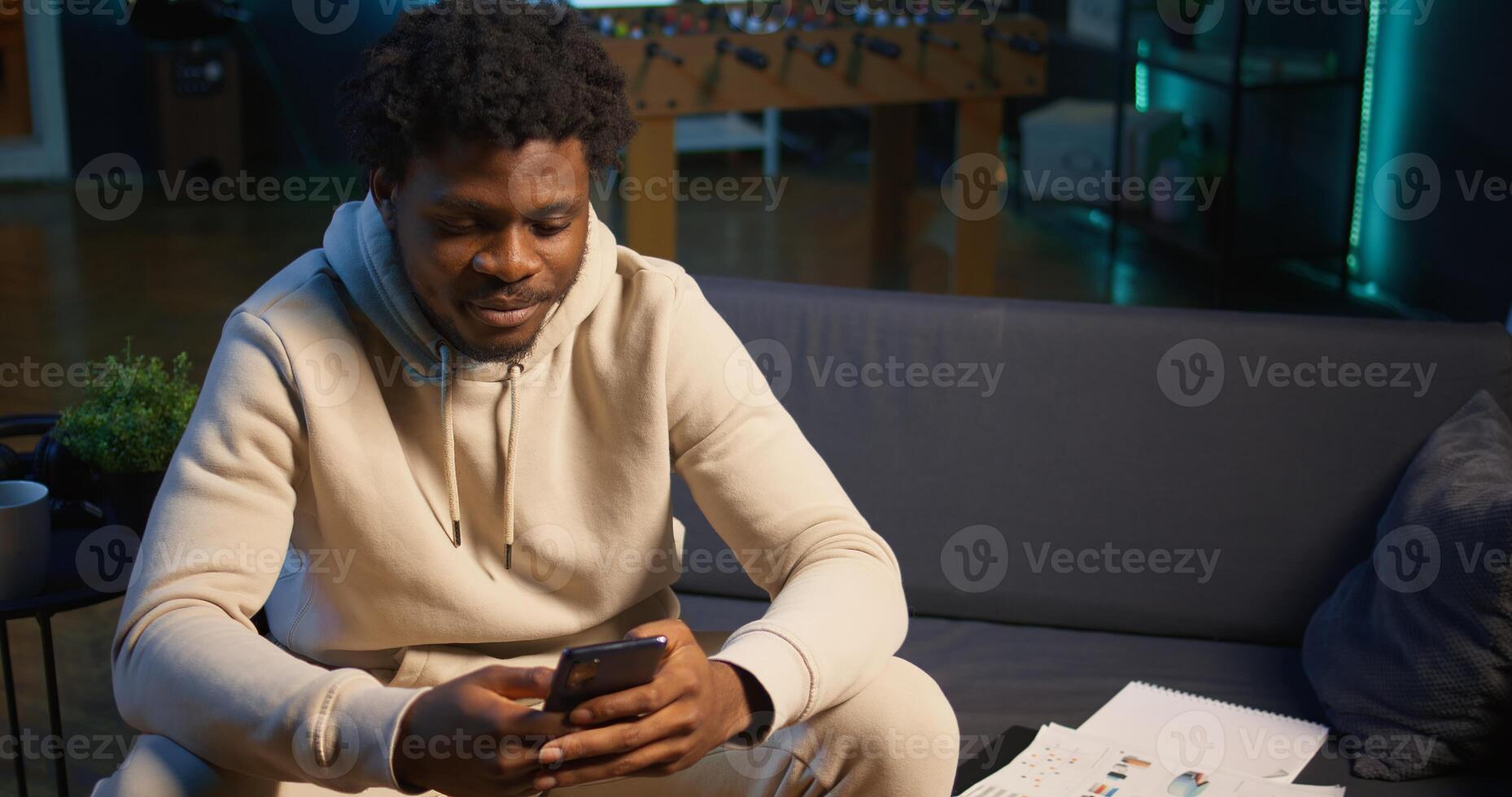 African American man on sofa texting mates in living room, entertained by social media and messages. Depiction of remote work breaks, digital communication and relaxation at home. Camera B. photo