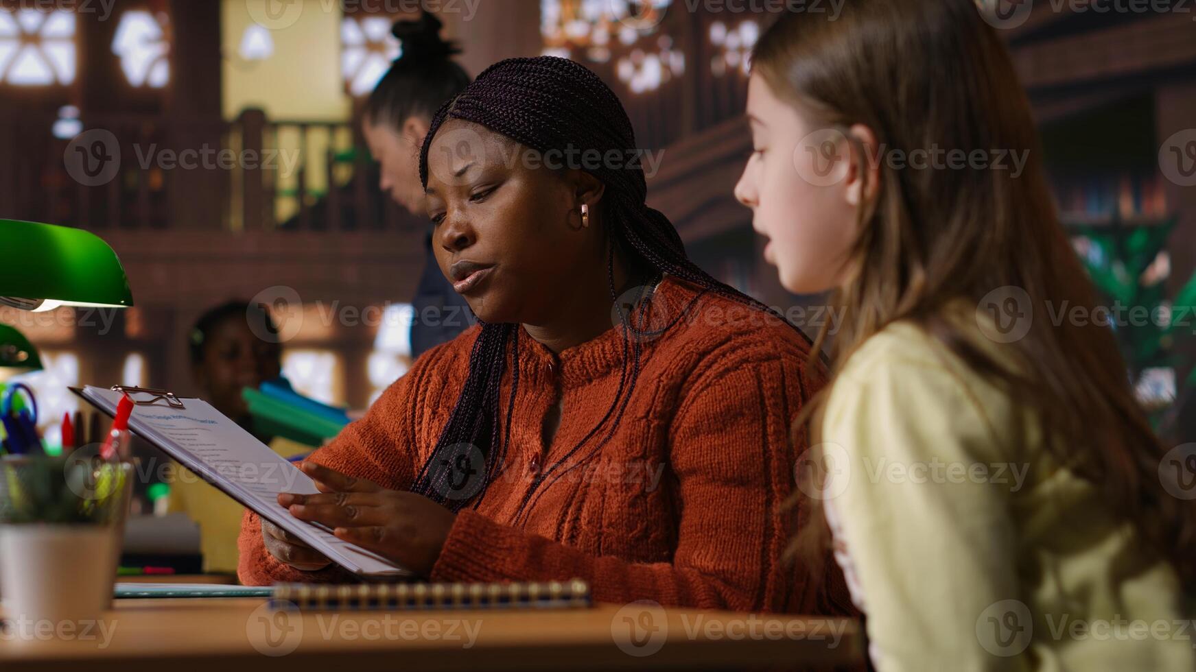 Black tutor mentoring schoolgirl in school library study desk, boosting her understanding of subject through personalized guidance. Professor hosts private lessons with pupil. Camera B. photo