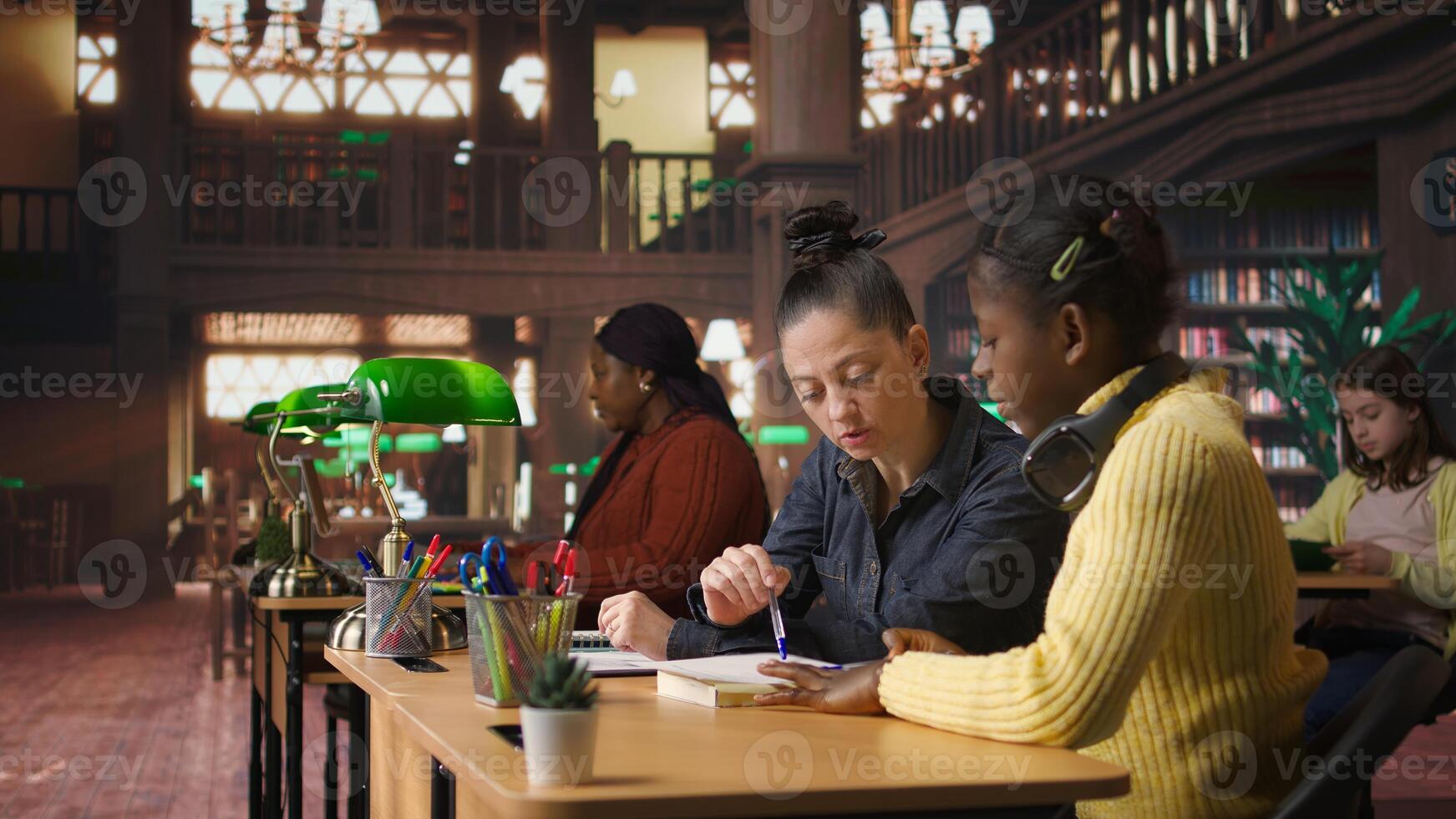 Caucasian tutor guiding elementary school girl at library desk, providing private lessons to aid in academic development with work books and notes. Teacher discussing with scholar. Camera A. photo