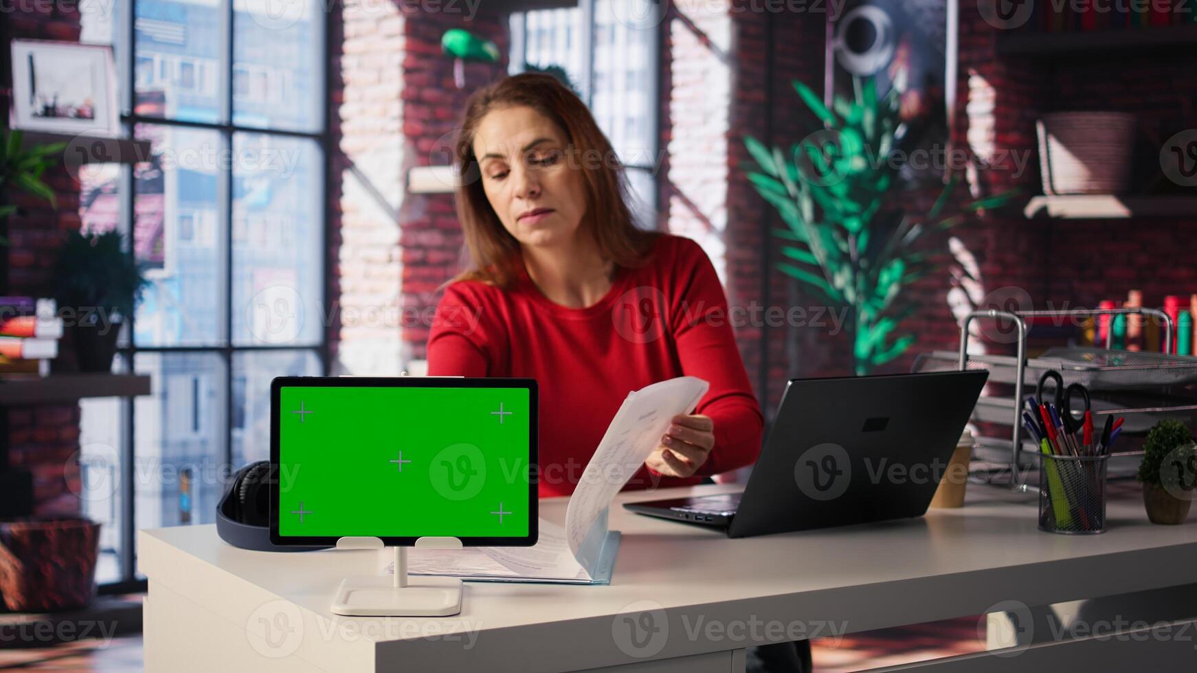 Freelancer does research by reading paper articles next to chroma key display, collecting information for new project. Woman multitasking and studying files with handwritten notes. Camera B. photo