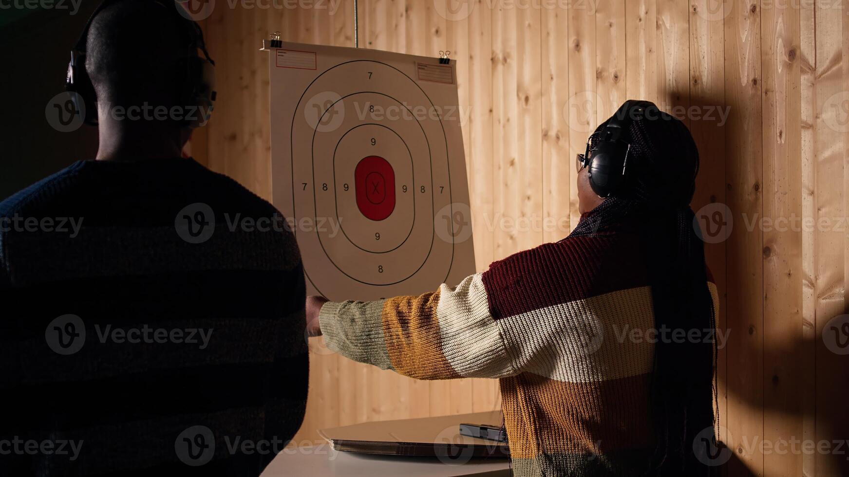 Woman in firing range training with pistol, looking at results on paper target. African american person doing shooting sport, sharpening skills and checking points scored on bullseye, camera A photo