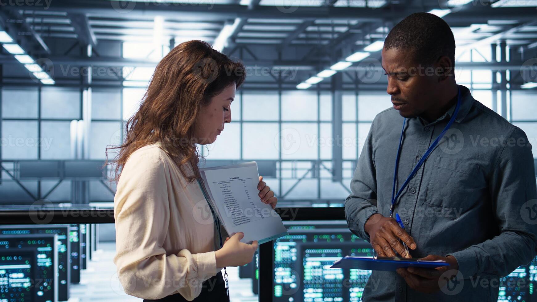 IT professionals looking over data center checkup checklist paperwork. African american man and colleague collaborating in server farm reading documentation files, camera B photo