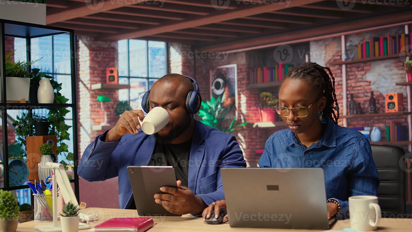 Black woman working while partner listens to music next to her, enjoying time at home together. Freelancer solving daily tasks on laptop, boyfriend scrolling web. Camera A. photo
