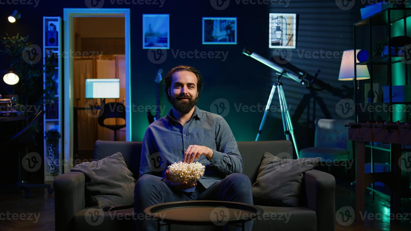 Man turning on TV using remote control, enjoying leisure time while munching on popcorn. Couch potato person in neon illuminated apartment eating salty snack, entertained by television, camera B photo