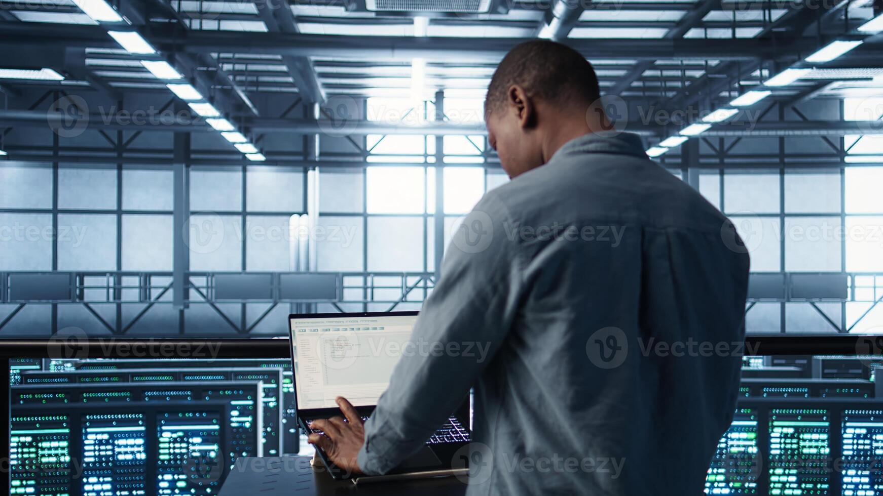 Computer scientist in server room coding on laptop, developing disaster recovery plans. African american man in data center testing failover scenarios using digital device, camera B photo