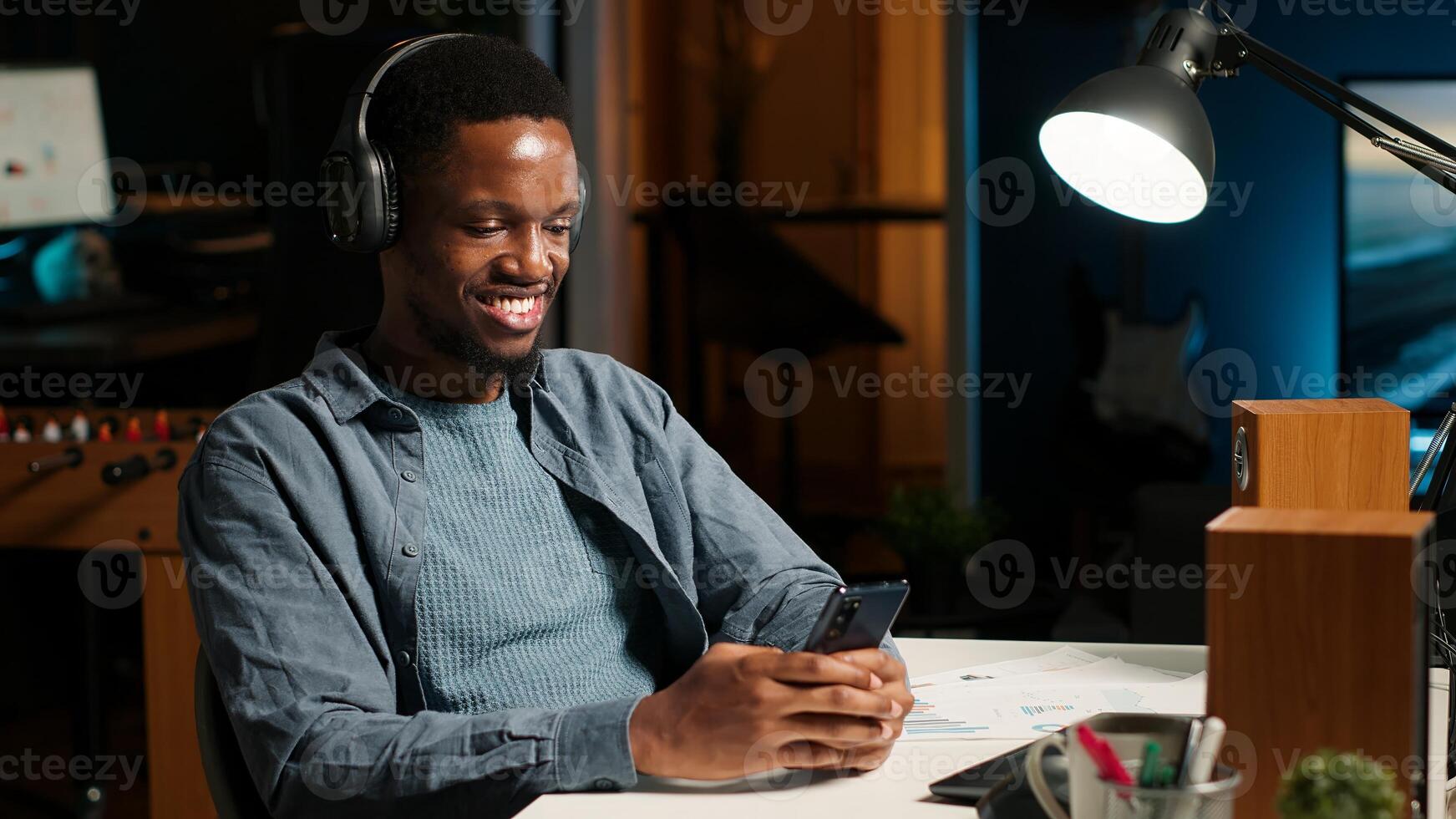 African american person takes break from work to answer texts at home, using smartphone to scroll social media apps. Young guy enjoying remote job, listening to music on device. Camera B. photo