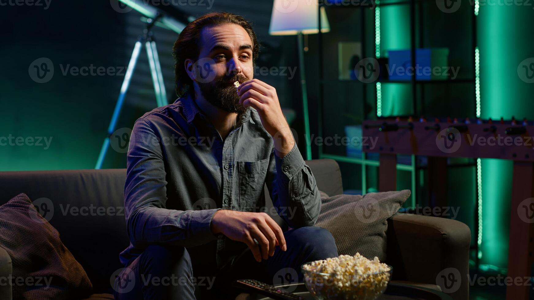 Man relaxing at home, stuffing mouth with popcorn while watching movies in apartment. Person eating snacks and streaming TV shows in neon lit living room, camera A photo