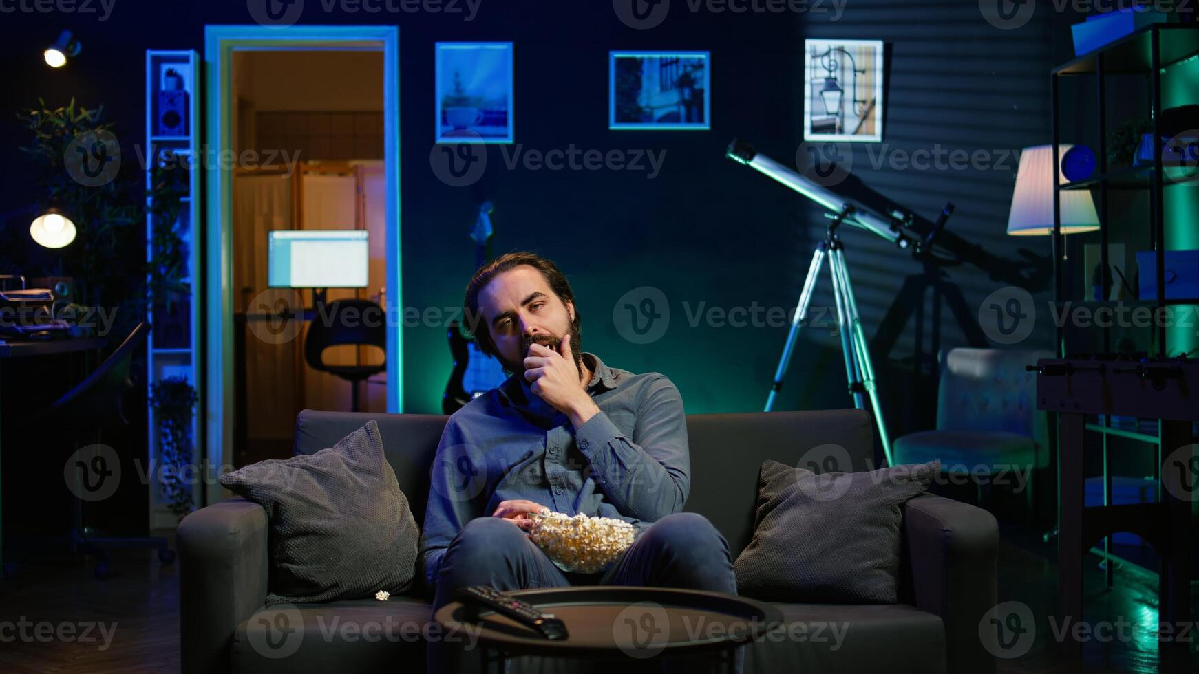 Fatigued man watching TV late at night and munching on popcorn, paying close attention to screen. Tired sedentary man sitting in pajamas all day consuming mindless entertainment, camera B photo
