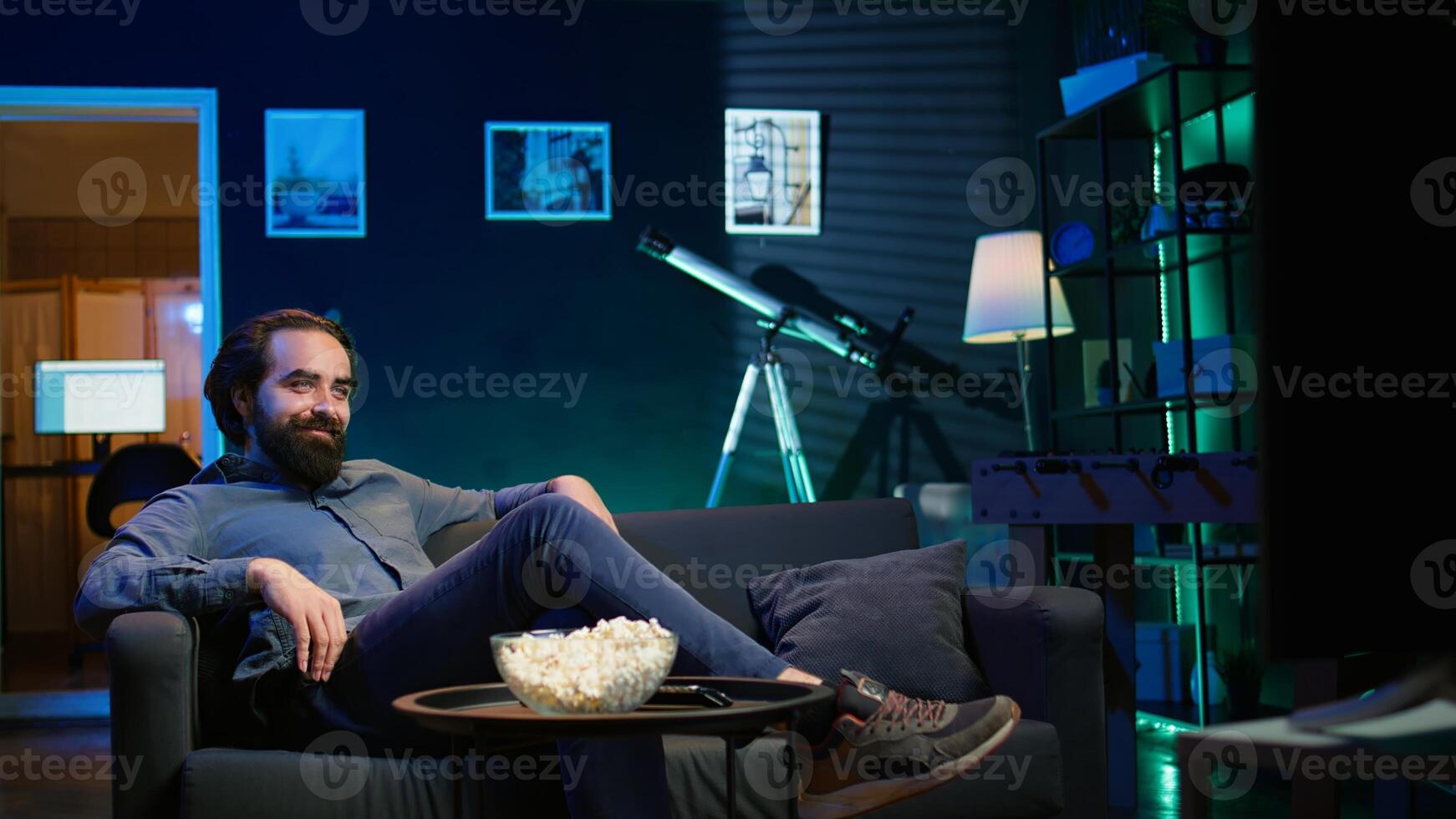 Amused man sitting on couch, watching funny comedy movie and eating popcorn. Cheerful person in apartment consuming snacks and laughing at hilarious joke in film scene, camera B photo