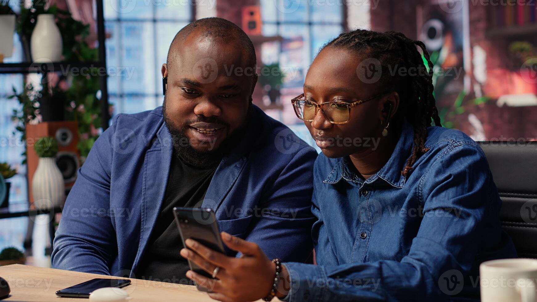 African american man and woman sit at desk discussing freelance strategies, researching marketing trends on phone to create helpful report. Couple doing teamwork. Camera B. photo