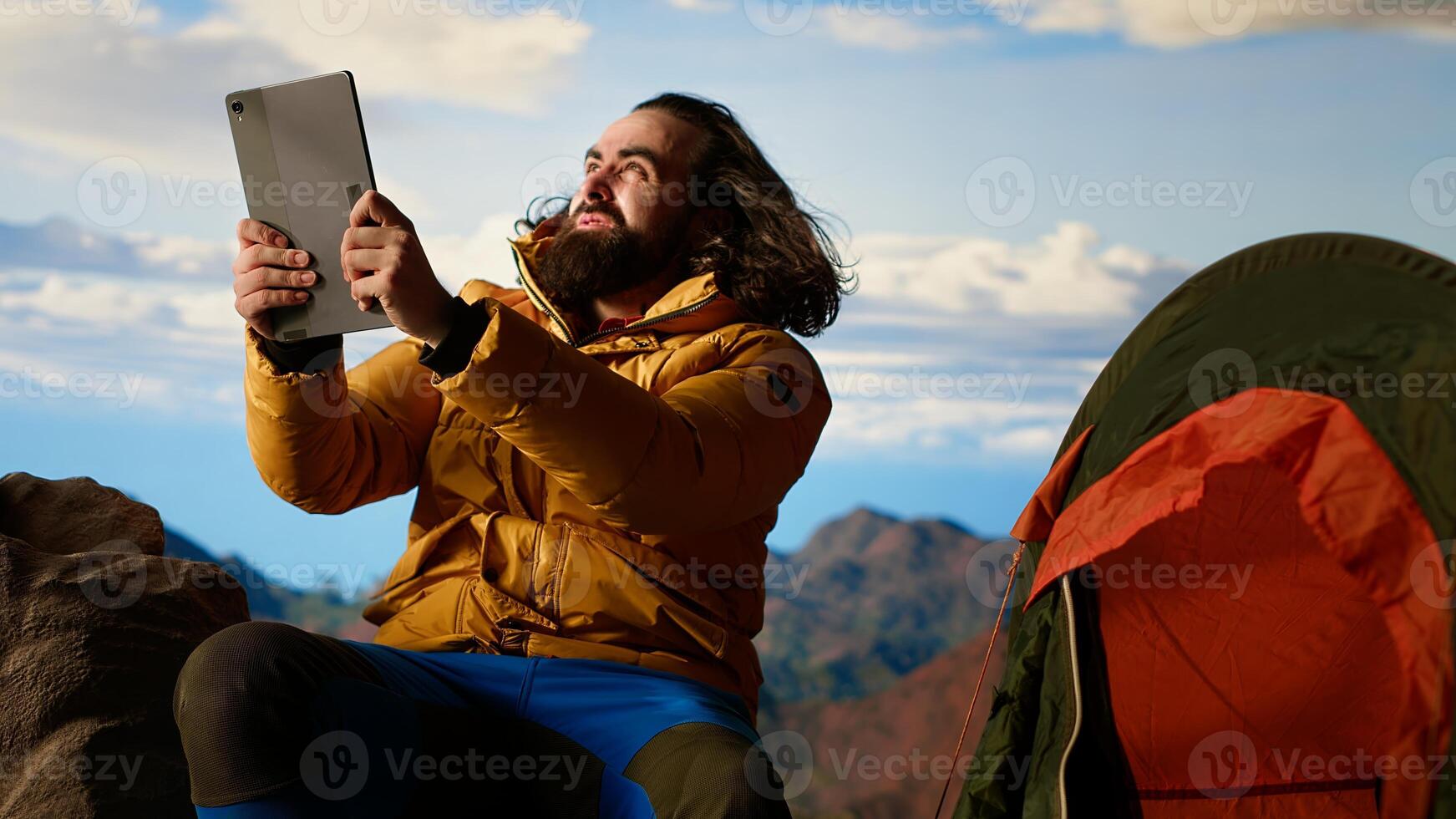 Reckless explorer struggling with wind and weak internet connection, camping on scenic rocky mountain peak. Hiker in danger with extreme cold weather. Camera B. photo
