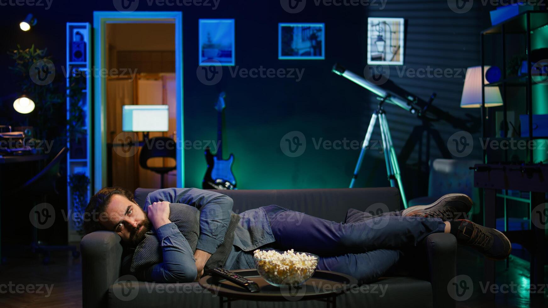 Man hugging pillow and watching TV, heartbroken by upsetting movie plot. Sad viewer laying on couch in RGB illuminated apartment, upset by depressing television show scene, camera B photo