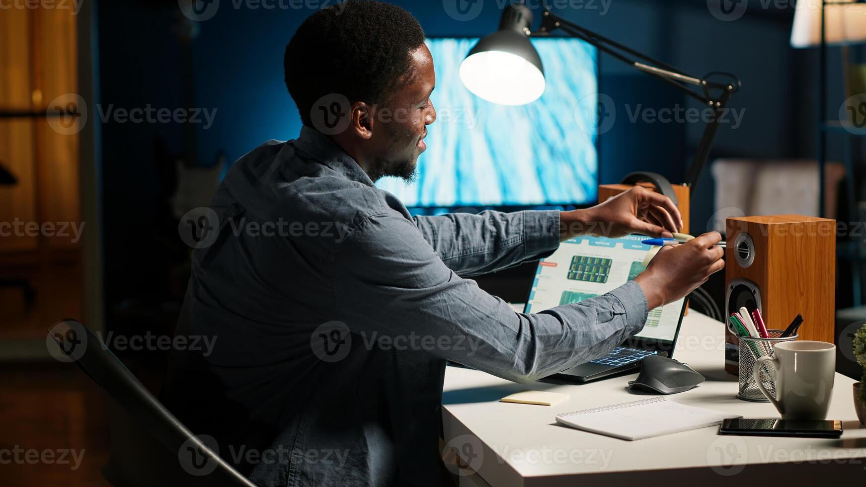 Black man organizing schedule with planner and putting reminder, creating adhesive note and browsing web. Modern productive guy writes memo for research. Camera B. photo