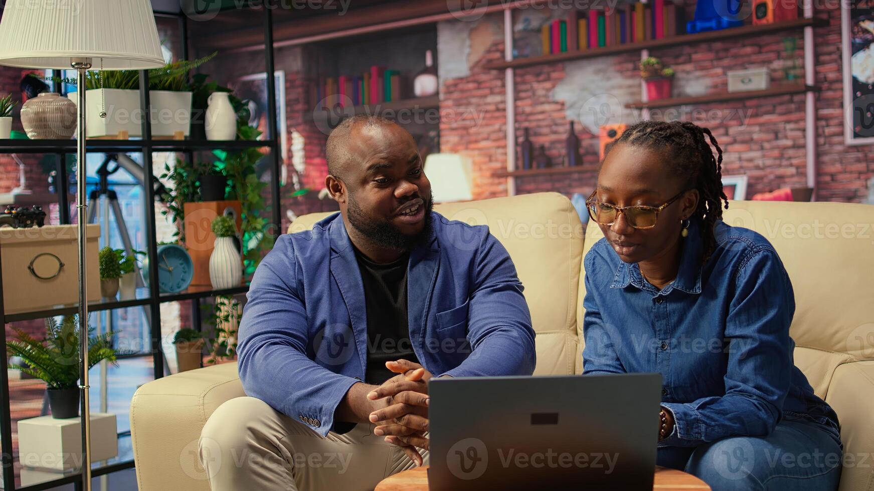 Black man and woman sit together in cozy apartment, discussing digital marketing strategy on laptop. Supporting each other in remote work and freelancing journey. Camera A. photo