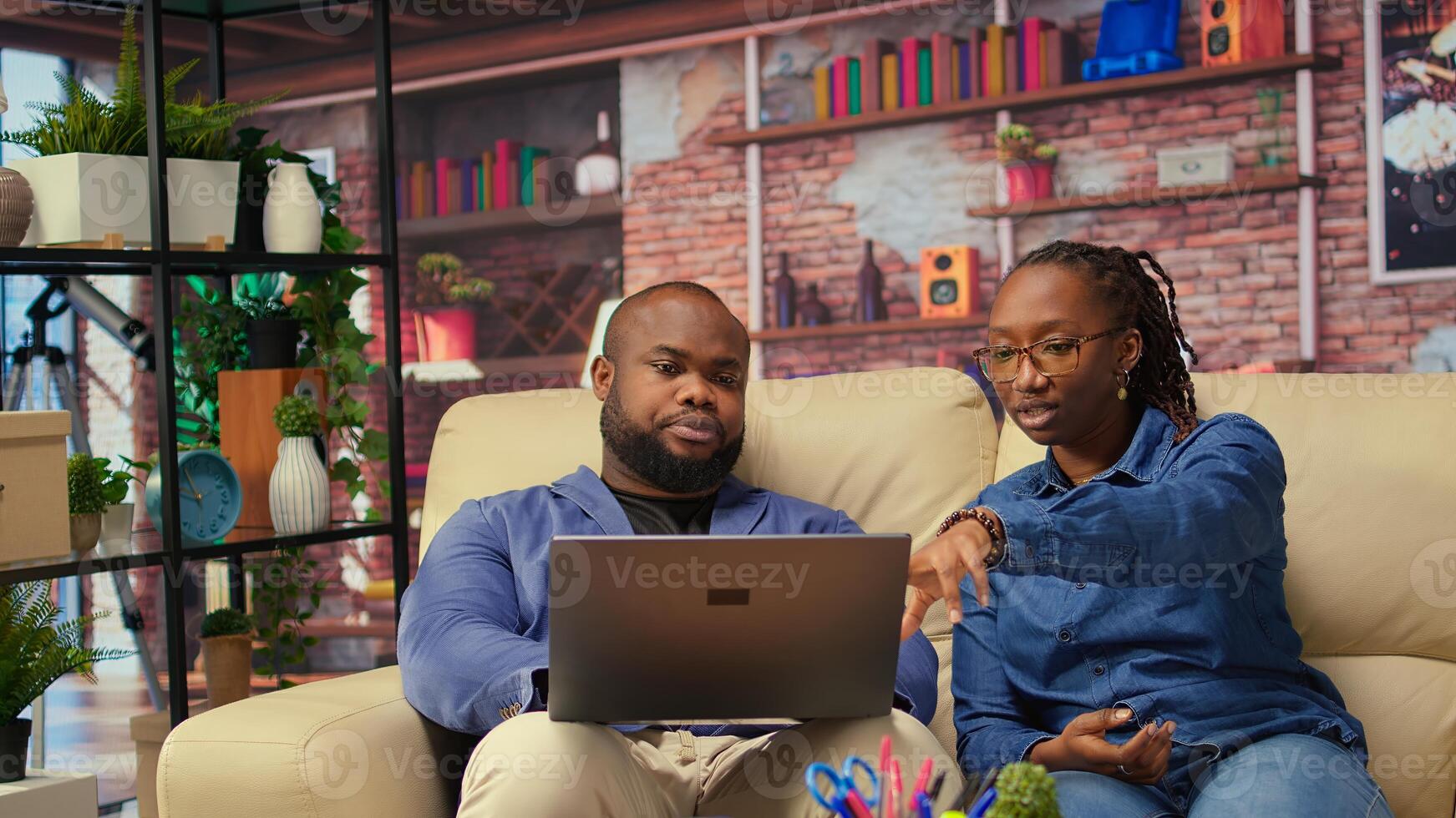 Teleworkers in living room looking at laptop screen and discussing about daily tasks, working from home. African american couple building career remotely. Camera A. photo