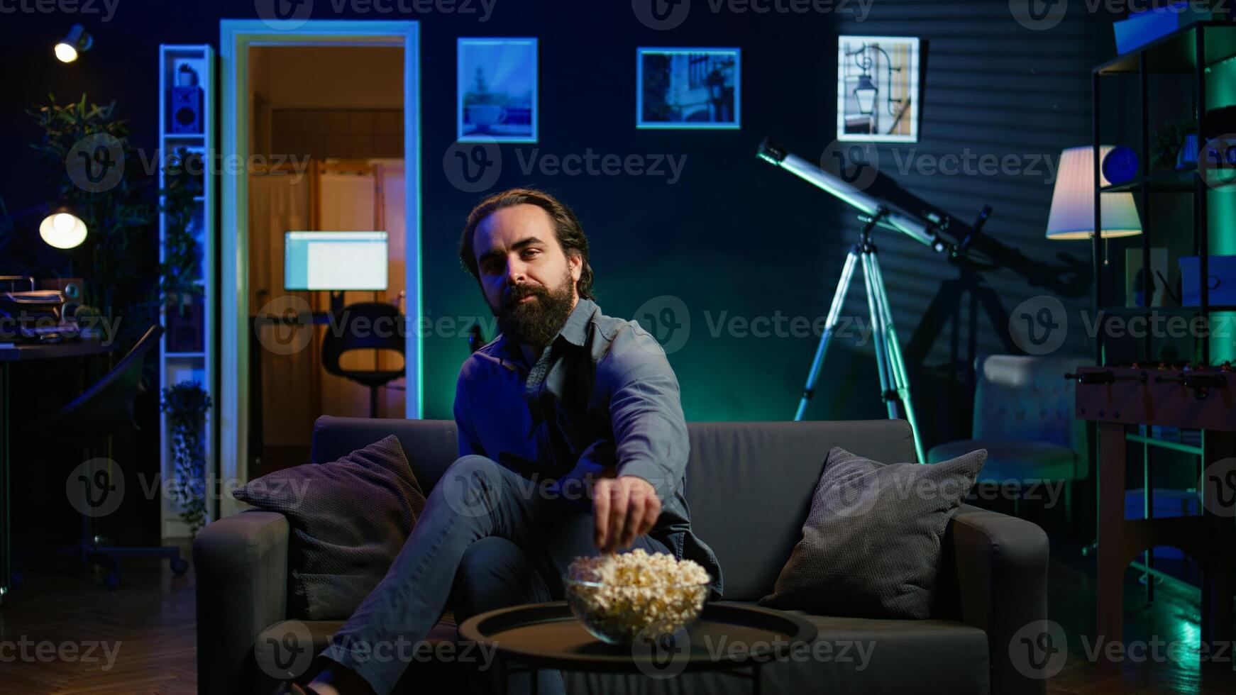Man taking seat on sofa, watching television and getting entertained in relaxed home. Person arriving in apartment sitting down on couch to enjoy movies, turning on TV, eats popcorn, camera B photo