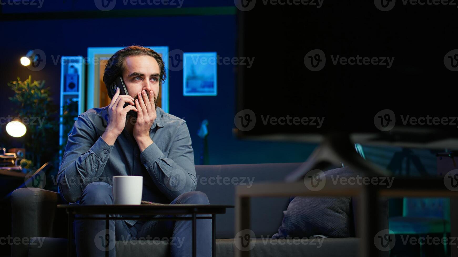 Man appalled by upsetting breaking news story on TV, on phone call with family member. Person at home scared by troubling events broadcasted on TV, calling friend using smartphone, camera B photo